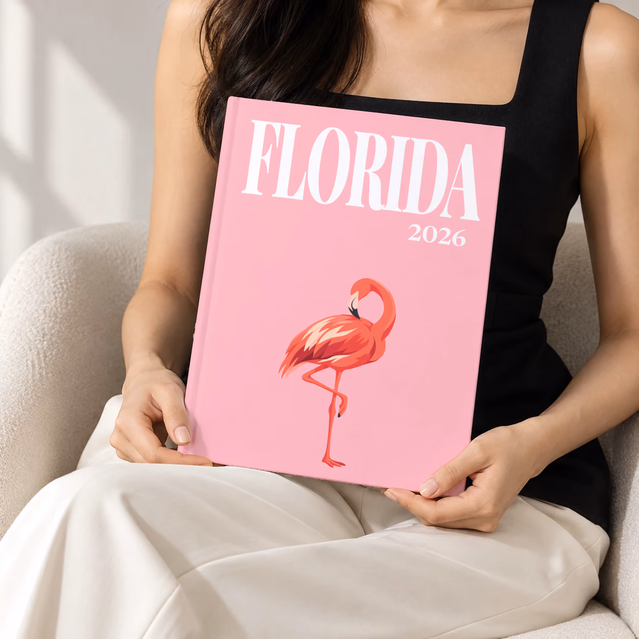 Florida hardcover photo book with the illustrated cover of a pink flamingo on pink, held upright in two hands by a woman in a black top, sitting in a cream boucle armchair