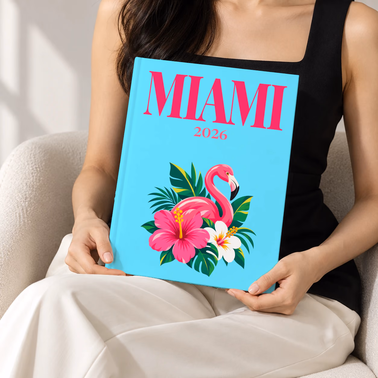 Florida hardcover photo book with the Miami illustrated cover of a pink flamingo with hibiscus flowers and palm leaves on aqua, held upright in two hands by a woman in a black top, sitting in a cream boucle armchair