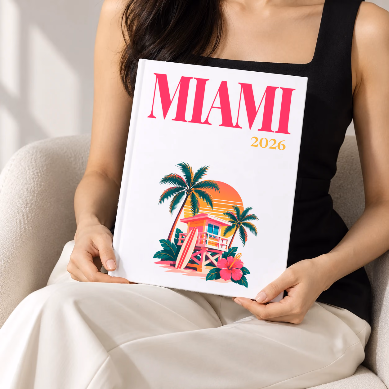 Florida hardcover photo book with the Miami illustrated cover of a pink Miami Beach lifeguard tower with palm trees at sunset on white, held upright in two hands by a woman in a black top, sitting in a cream boucle armchair