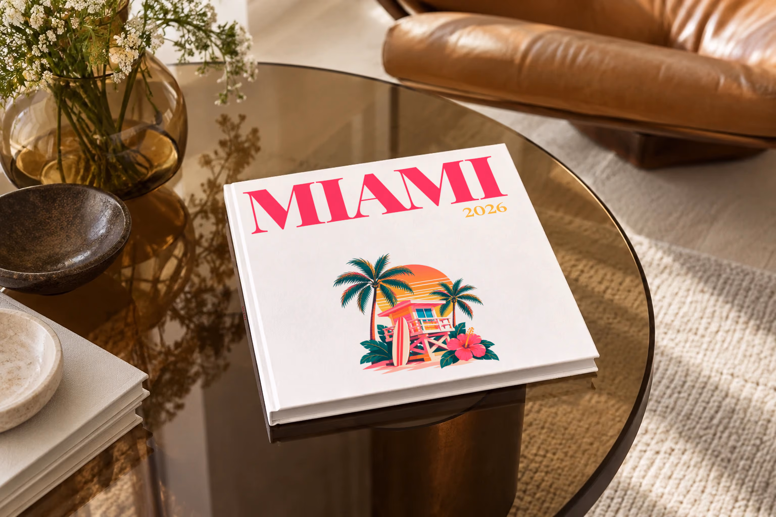 Florida square hardcover photo book with the Miami illustrated cover of a pink Miami Beach lifeguard tower with palm trees at sunset on white, on a smoked glass coffee table beside a leather armchair in warm afternoon light