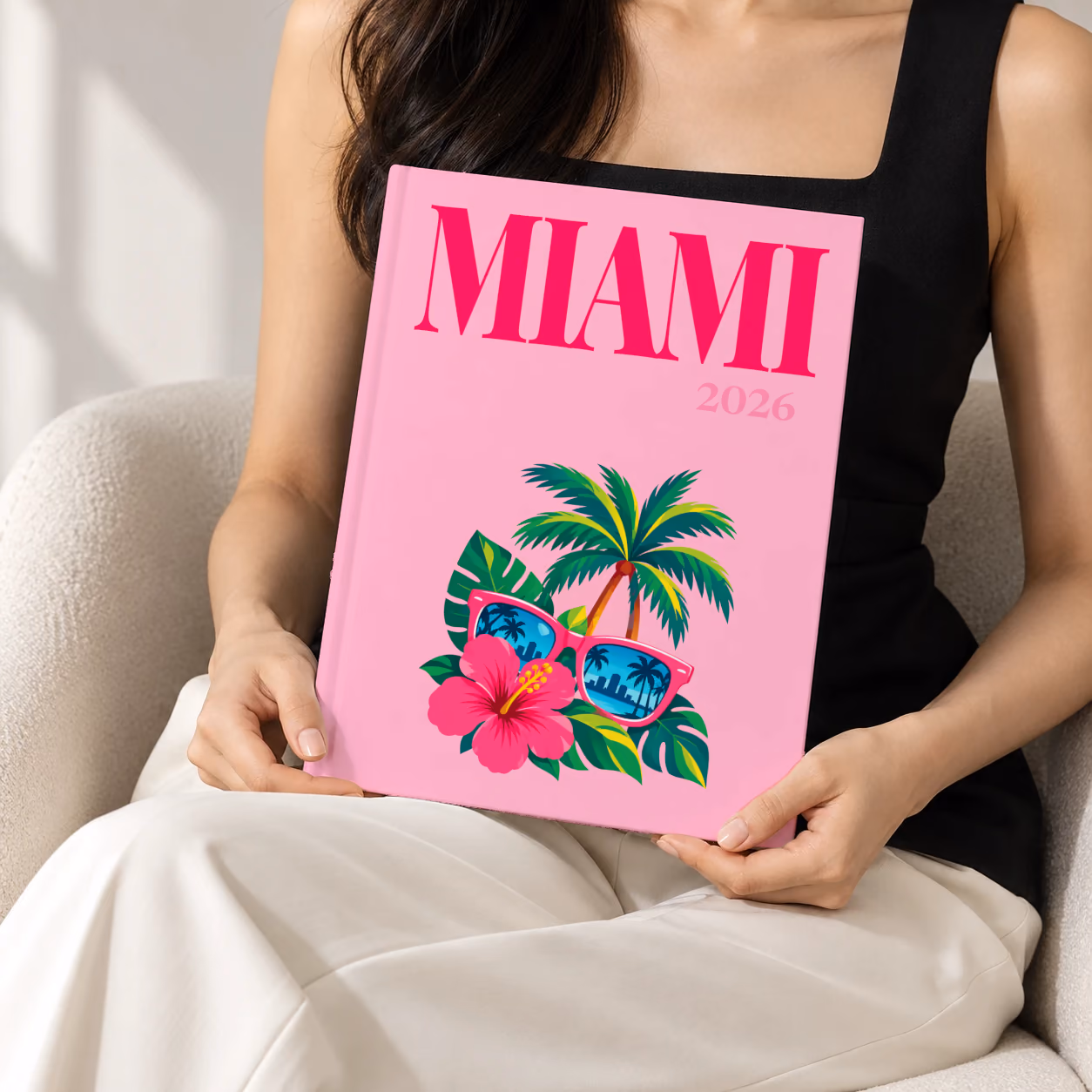 Florida hardcover photo book with the Miami illustrated cover of pink sunglasses reflecting palm trees, with hibiscus, on pink, held upright in two hands by a woman in a black top, sitting in a cream boucle armchair