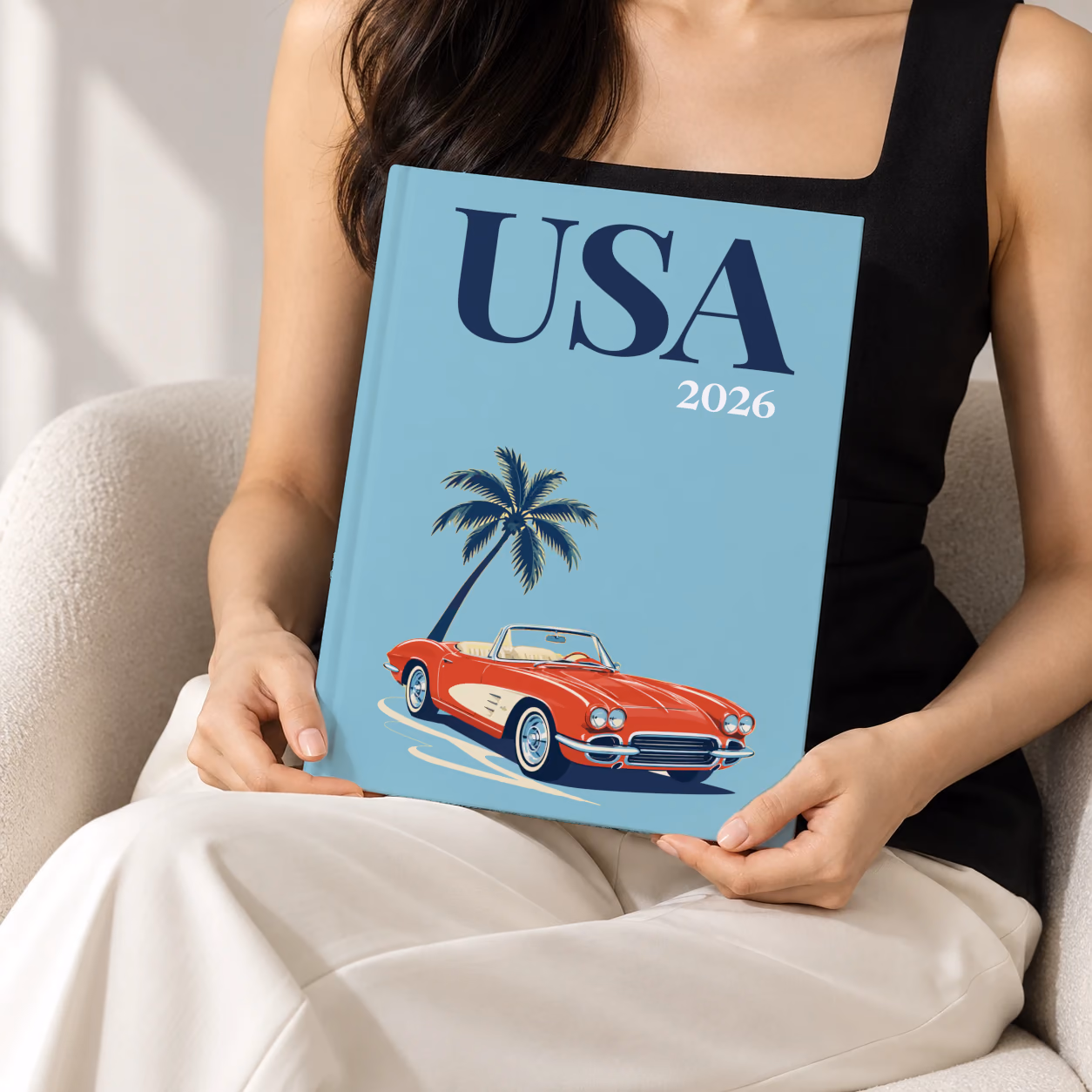 Florida hardcover photo book with the USA illustrated cover of a red Corvette and a palm tree on dusty blue, held upright in two hands by a woman in a black top, sitting in a cream boucle armchair