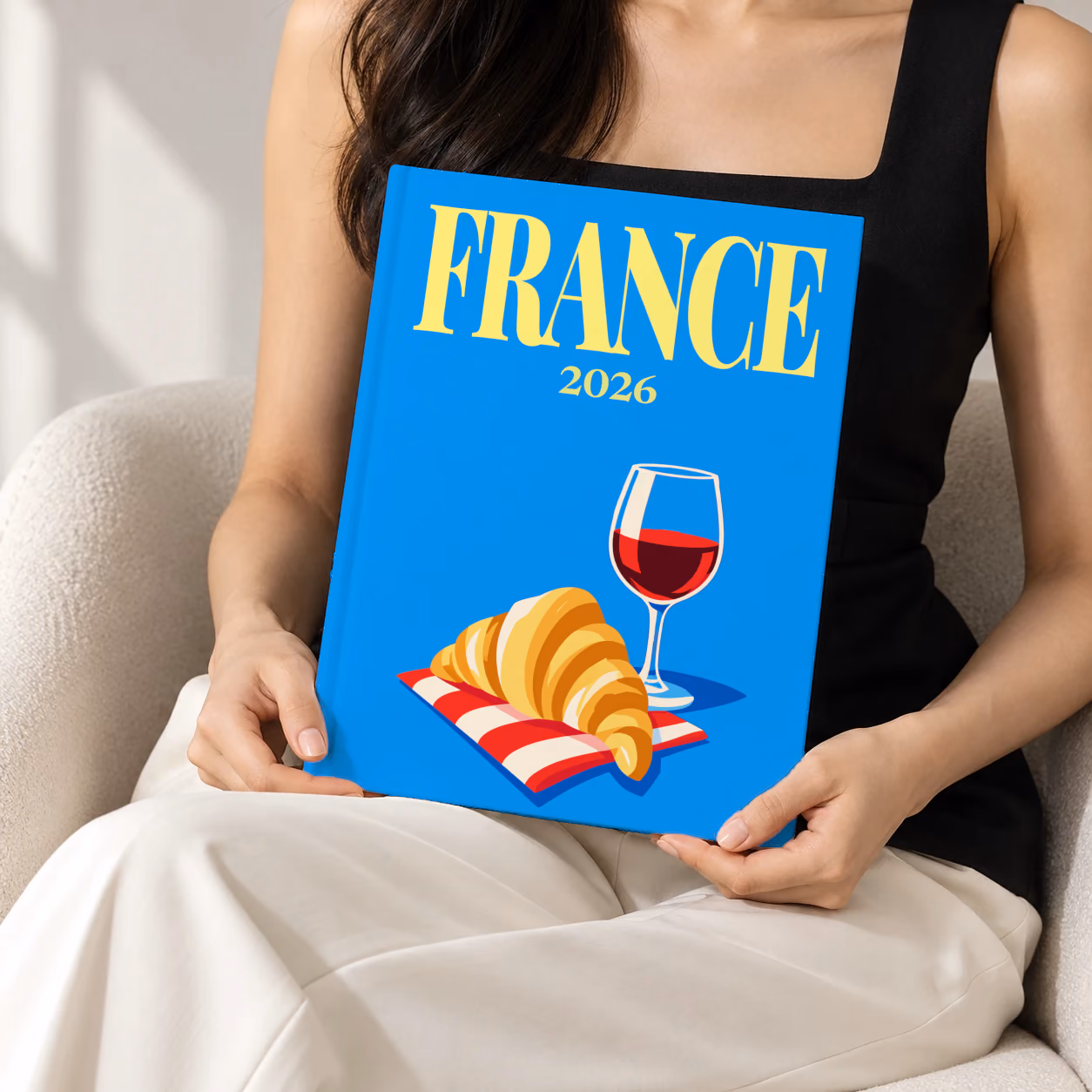 France hardcover photo book with the illustrated cover of a croissant and a glass of red wine on bright blue, held upright in two hands by a woman in a black top, sitting in a cream boucle armchair