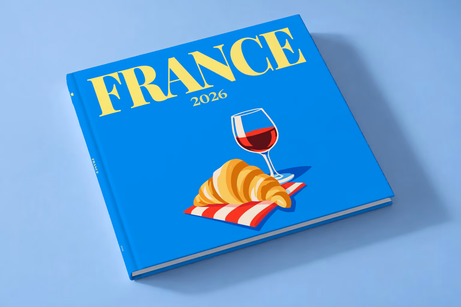 France square hardcover photo book with the illustrated cover of a croissant and a glass of red wine on bright blue, lying on a pale blue studio backdrop