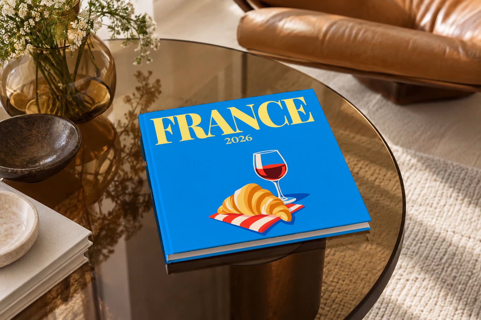 France square hardcover photo book with the illustrated cover of a croissant and a glass of red wine on bright blue, on a smoked glass coffee table beside a leather armchair in warm afternoon light