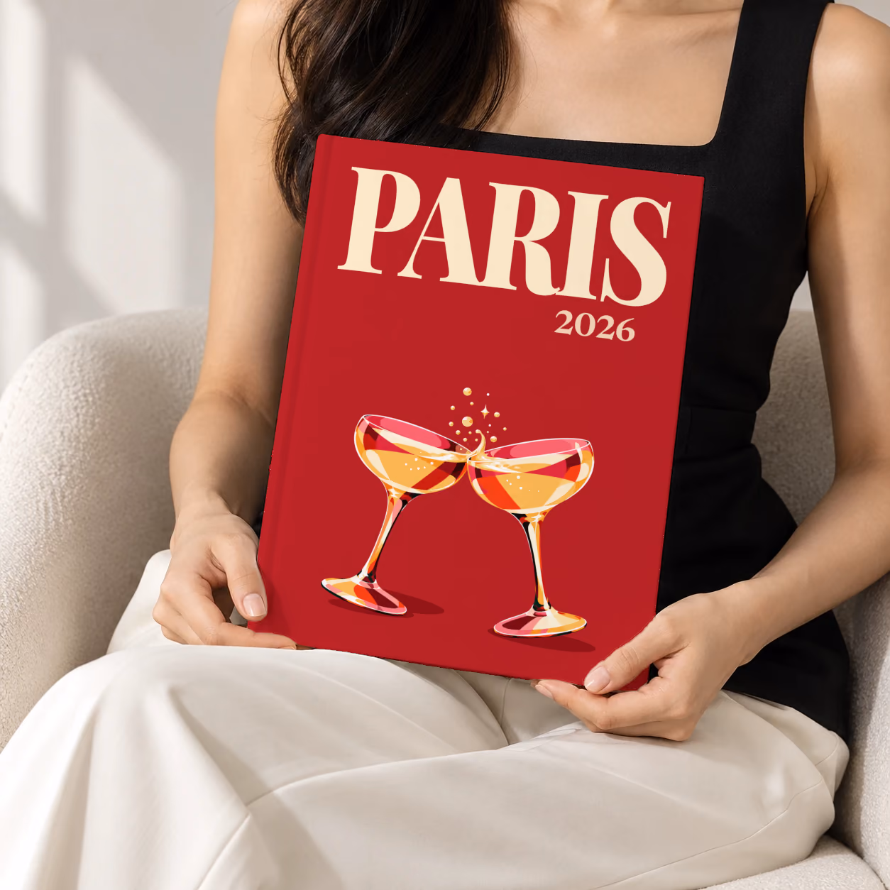 France hardcover photo book with the Paris illustrated cover of two champagne coupes on deep red, held upright in two hands by a woman in a black top, sitting in a cream boucle armchair