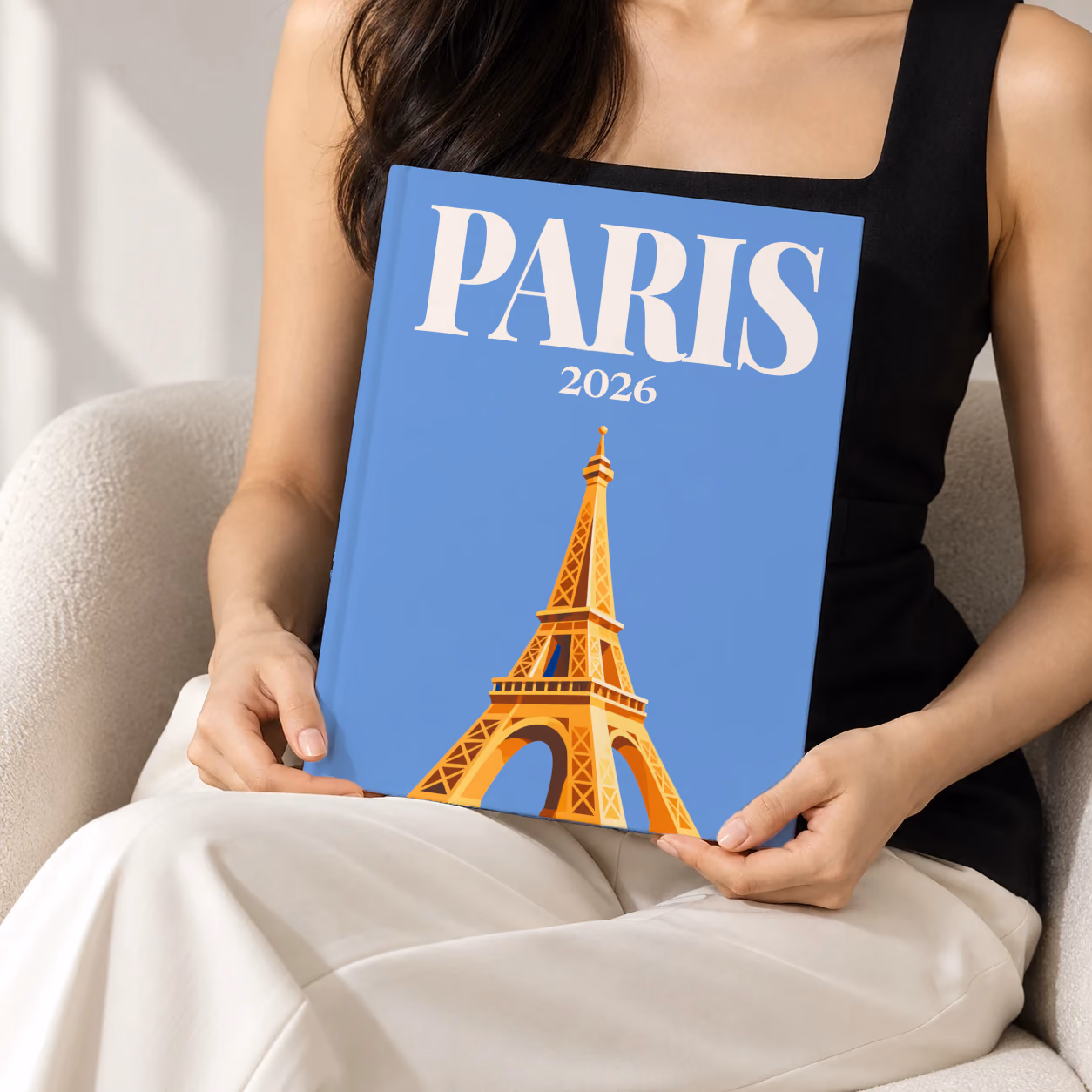 France hardcover photo book with the Paris illustrated cover of a gold Eiffel Tower on blue, held upright in two hands by a woman in a black top, sitting in a cream boucle armchair