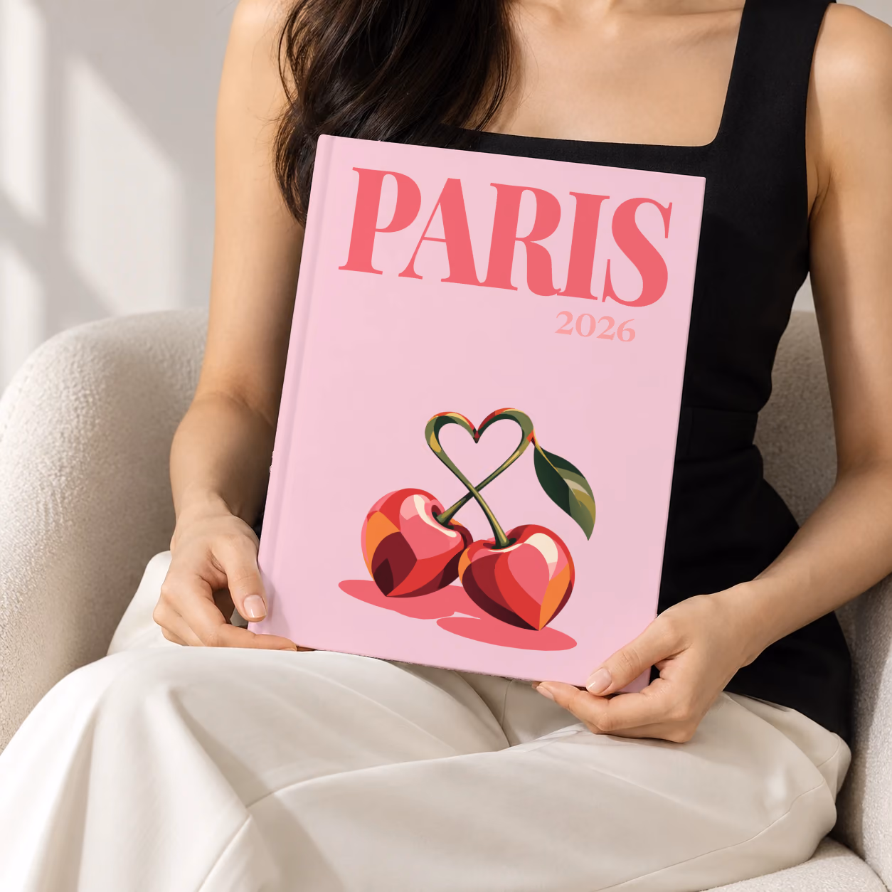 France hardcover photo book with the Paris illustrated cover of cherries with heart-shaped stems on pale pink, held upright in two hands by a woman in a black top, sitting in a cream boucle armchair