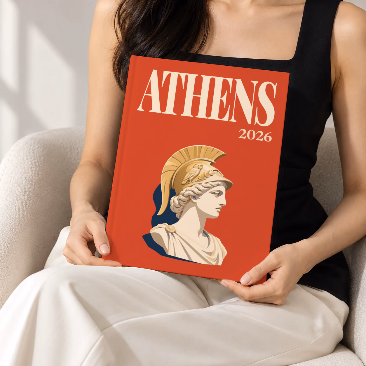Greece hardcover photo book with the Athens illustrated cover of a bust of Athena in a helmet on red, held upright in two hands by a woman in a black top, sitting in a cream boucle armchair