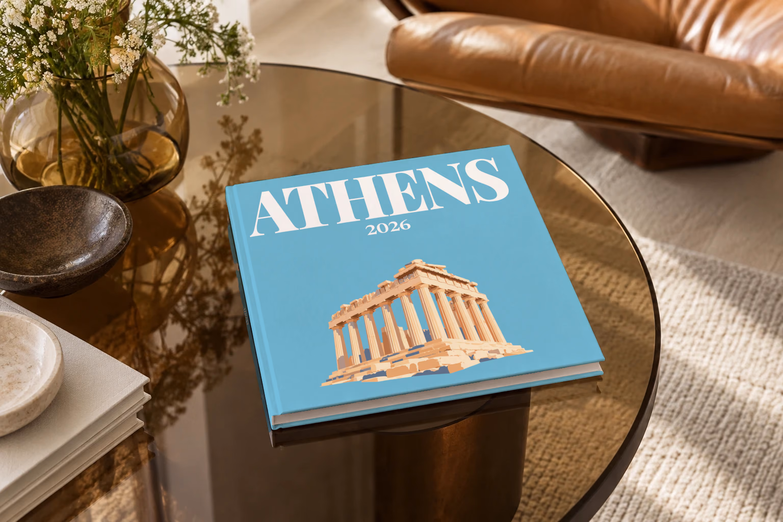 Greece square hardcover photo book with the Athens illustrated cover of the Parthenon on blue, on a smoked glass coffee table beside a leather armchair in warm afternoon light