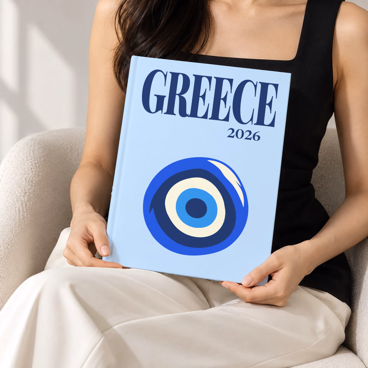 Greece hardcover photo book with the illustrated cover of an evil eye on pale blue, held upright in two hands by a woman in a black top, sitting in a cream boucle armchair