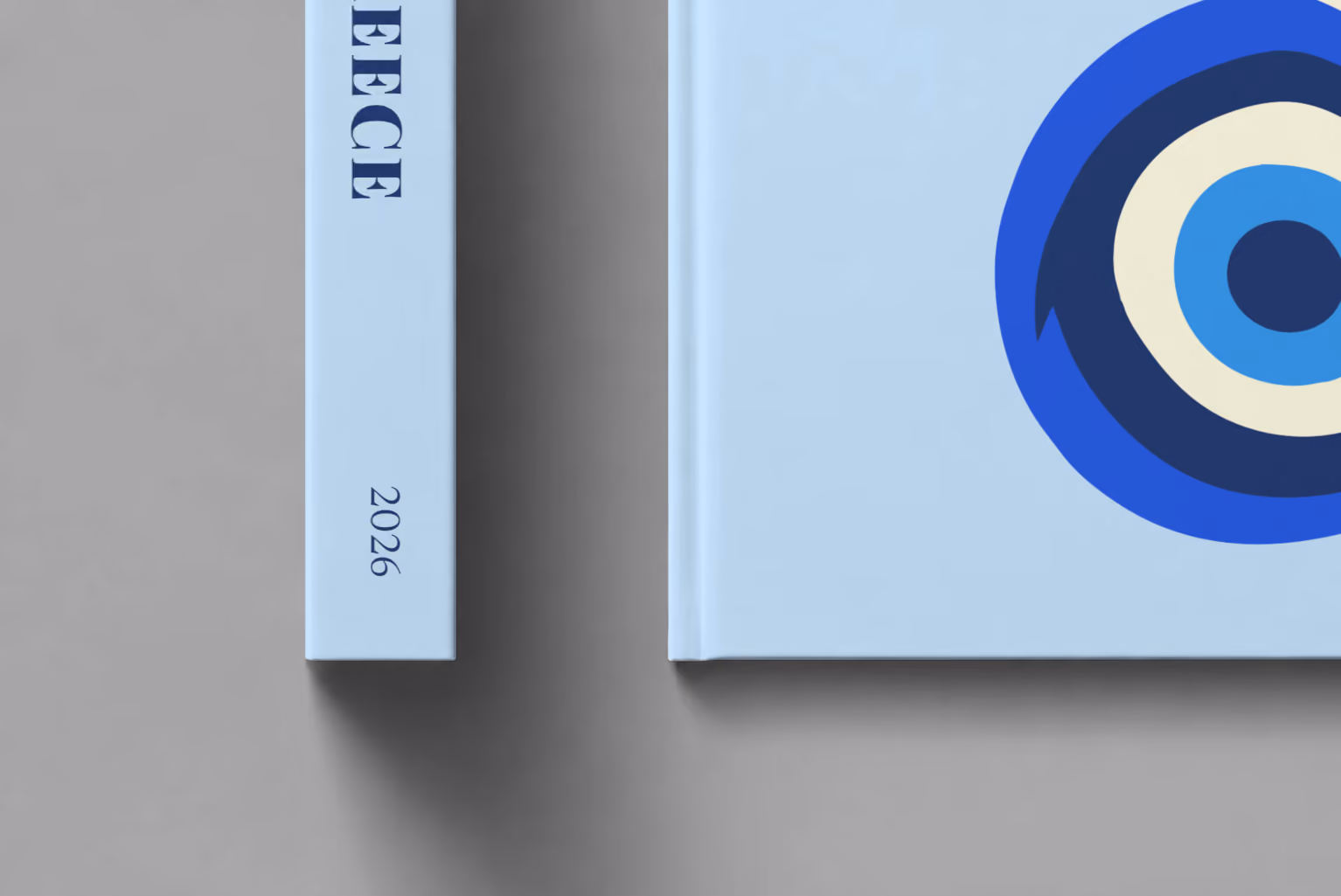 Greece hardcover photo book with the illustrated cover of an evil eye on pale blue, shown close up beside its spine on a pale grey studio surface