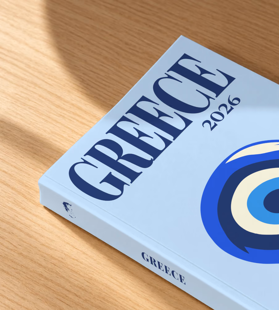 Greece softcover photo book with the illustrated cover of an evil eye on pale blue, lying on an oak desk in dappled afternoon sunlight