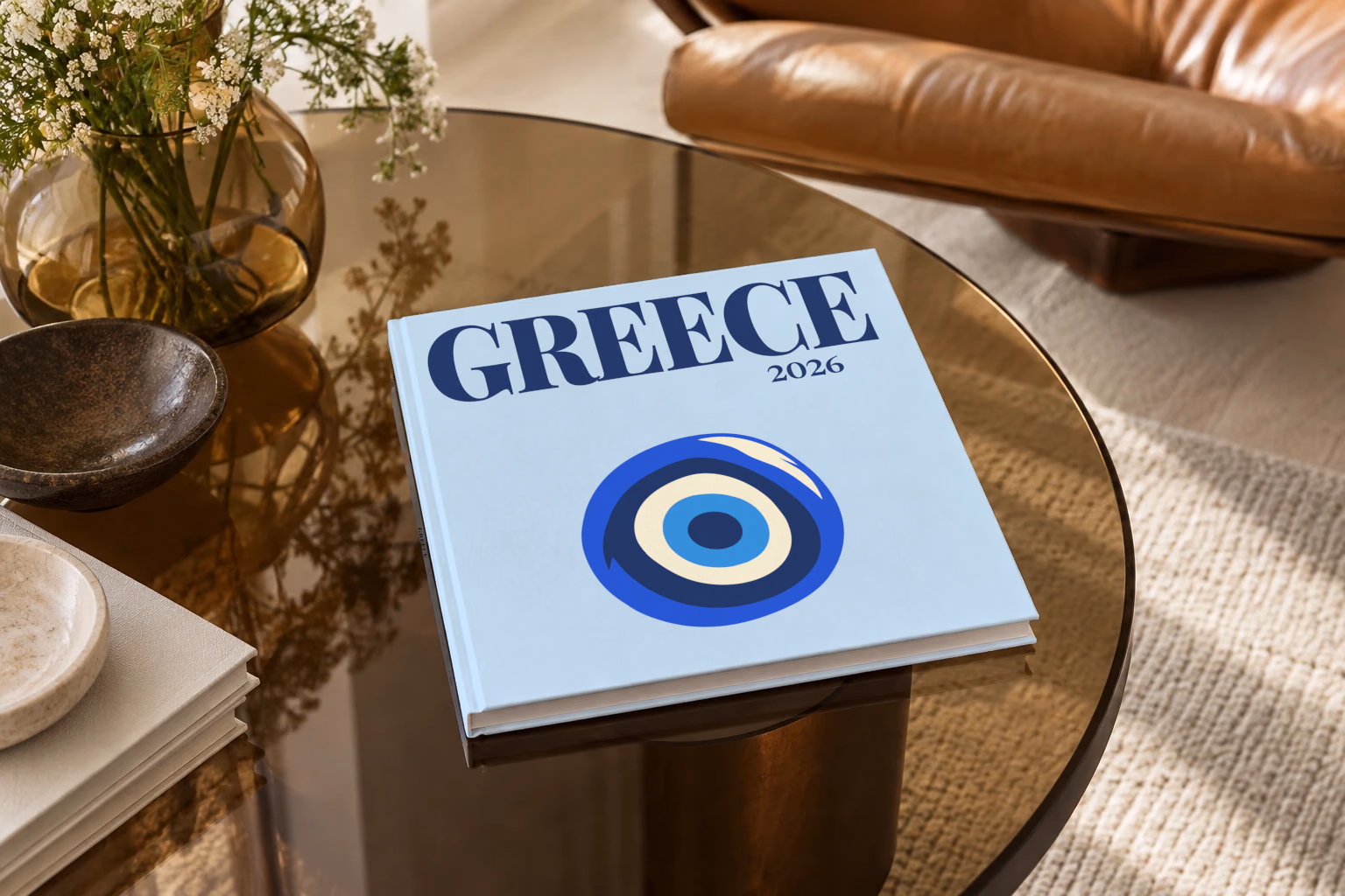 Greece square hardcover photo book with the illustrated cover of an evil eye on pale blue, on a smoked glass coffee table beside a leather armchair in warm afternoon light