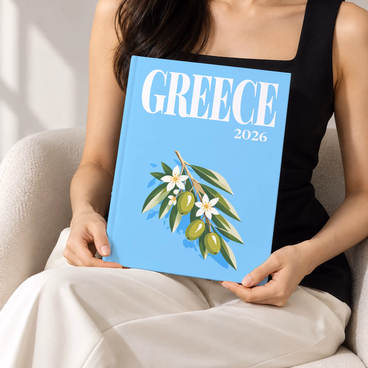 Greece hardcover photo book with the illustrated cover of an olive branch with blossom on sky blue, held upright in two hands by a woman in a black top, sitting in a cream boucle armchair