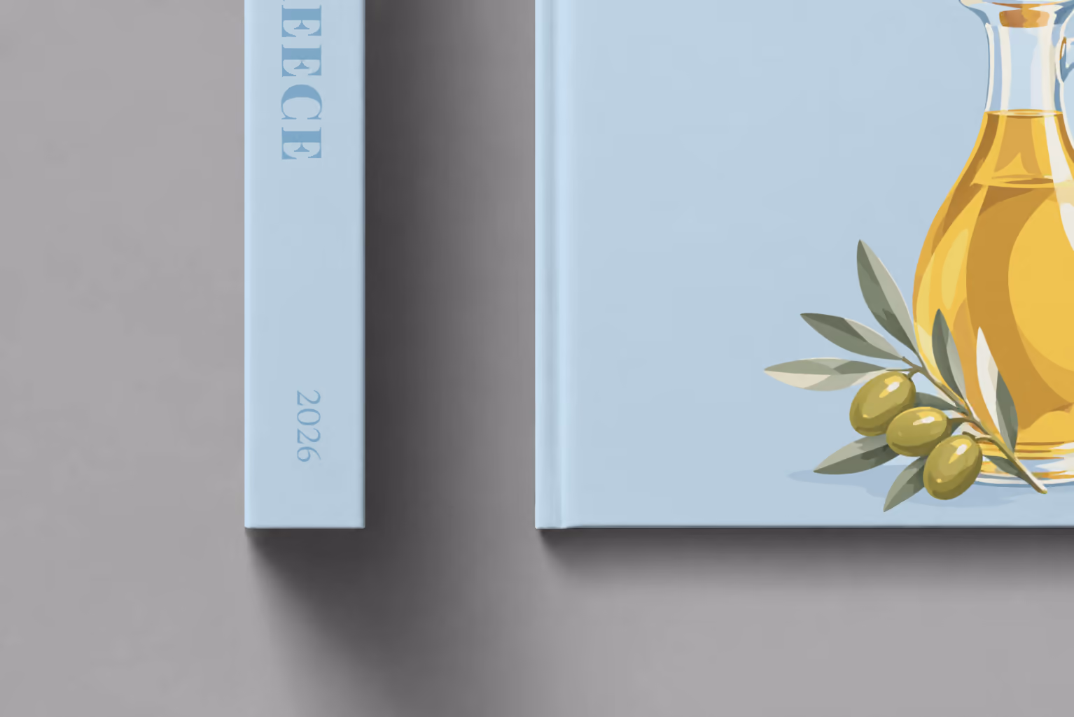 Greece hardcover photo book with the illustrated cover of a bottle of olive oil and olives on pale blue, shown close up beside its spine on a pale grey studio surface