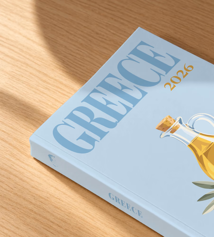 Greece softcover photo book with the illustrated cover of a bottle of olive oil and olives on pale blue, lying on an oak desk in dappled afternoon sunlight