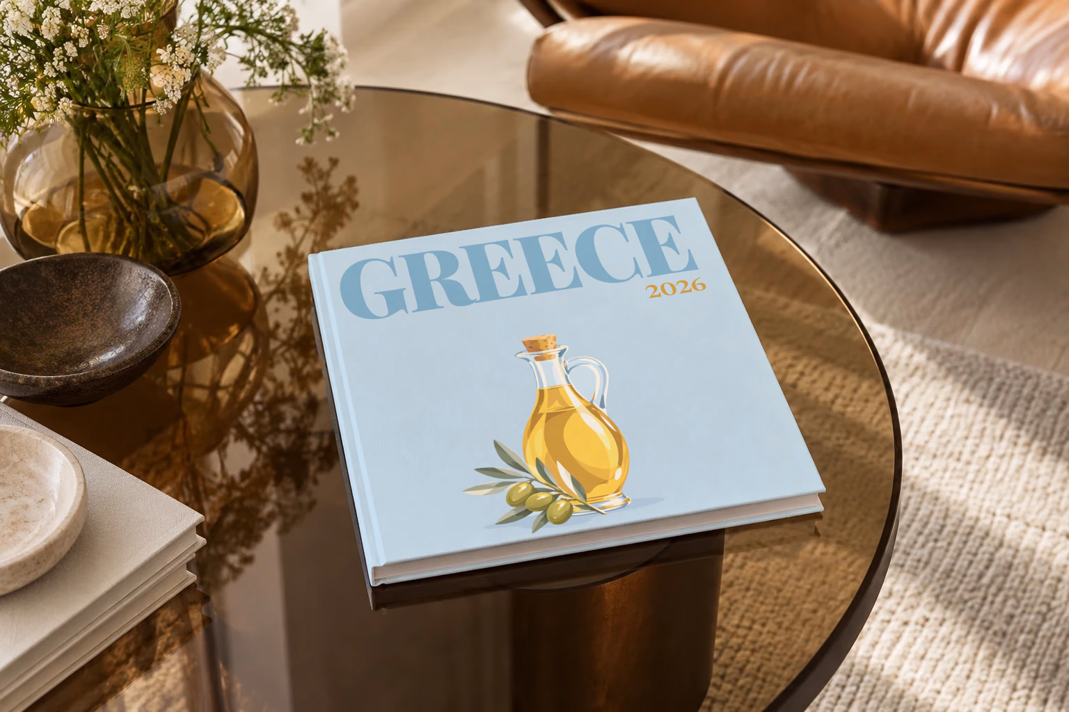 Greece square hardcover photo book with the illustrated cover of a bottle of olive oil and olives on pale blue, on a smoked glass coffee table beside a leather armchair in warm afternoon light
