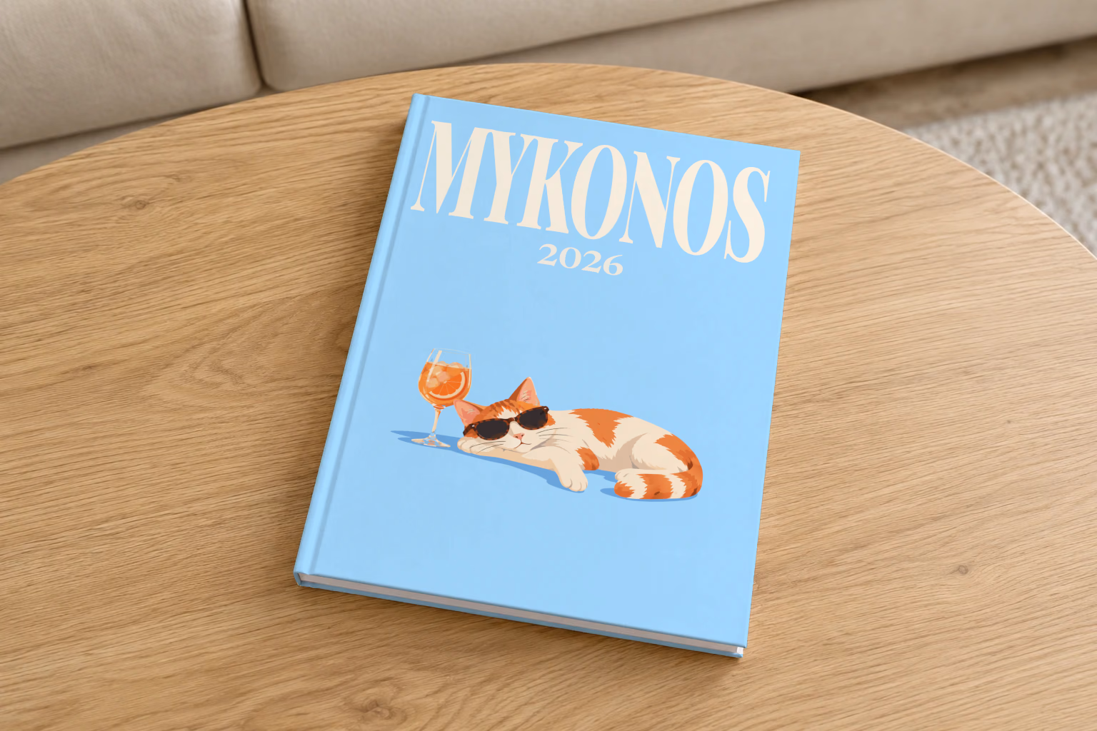 Greece hardcover photo book with the Mykonos illustrated cover of a cat in sunglasses beside a spritz on sky blue, on a round oak coffee table in a bright living room