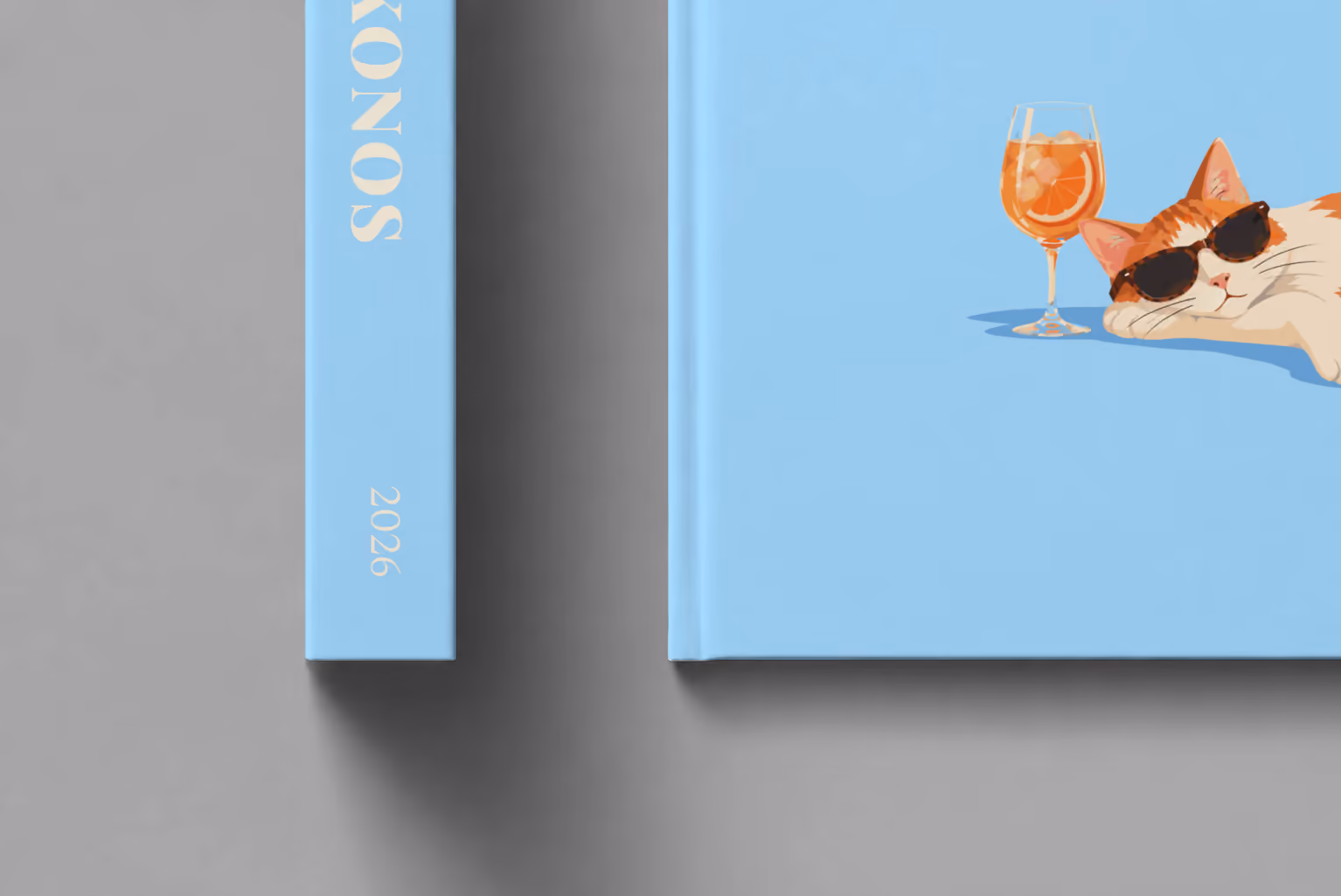 Greece hardcover photo book with the Mykonos illustrated cover of a cat in sunglasses beside a spritz on sky blue, shown close up beside its spine on a pale grey studio surface