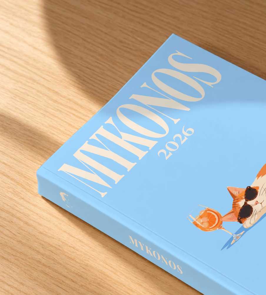 Greece softcover photo book with the Mykonos illustrated cover of a cat in sunglasses beside a spritz on sky blue, lying on an oak desk in dappled afternoon sunlight