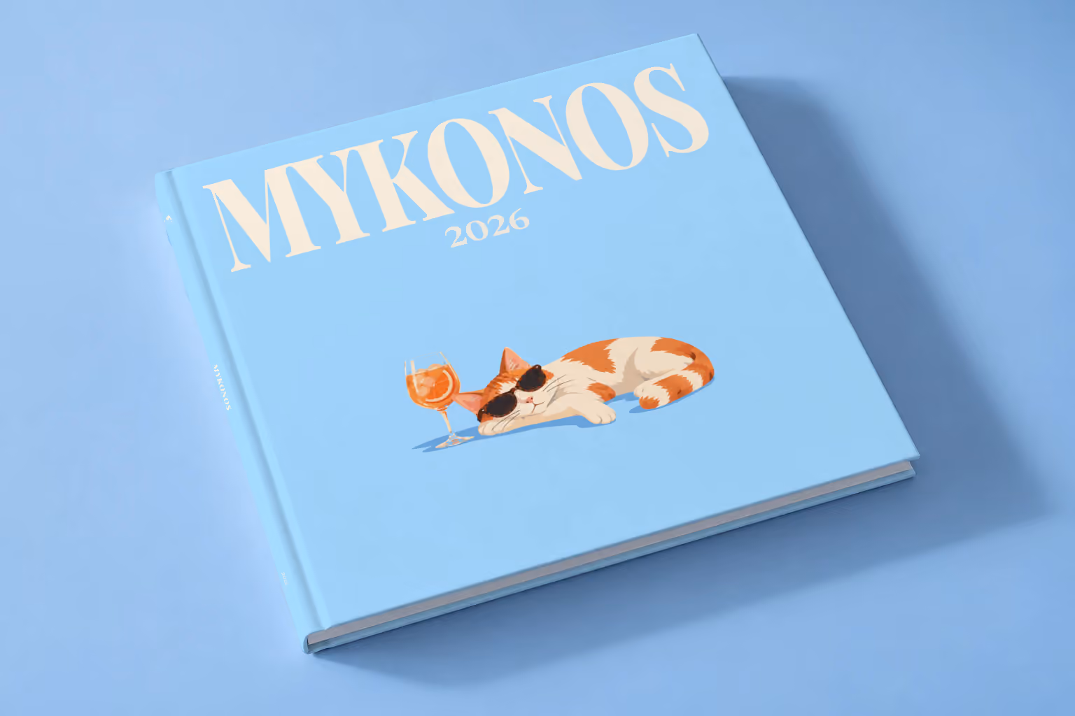 Greece square hardcover photo book with the Mykonos illustrated cover of a cat in sunglasses beside a spritz on sky blue, lying on a pale blue studio backdrop