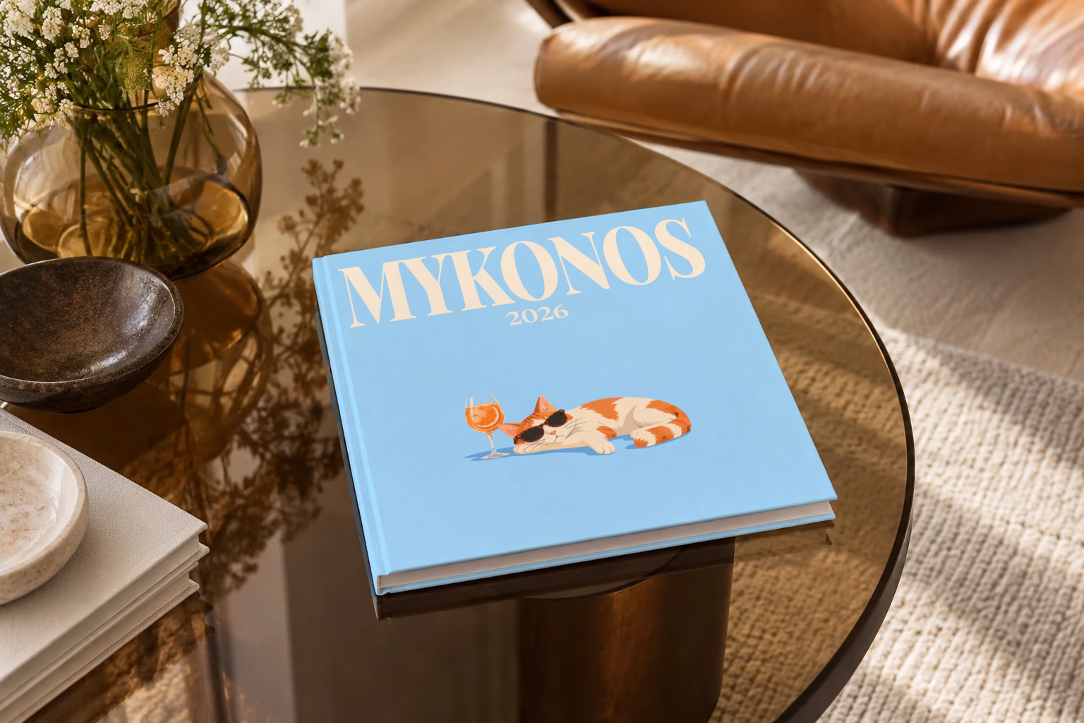 Greece square hardcover photo book with the Mykonos illustrated cover of a cat in sunglasses beside a spritz on sky blue, on a smoked glass coffee table beside a leather armchair in warm afternoon light