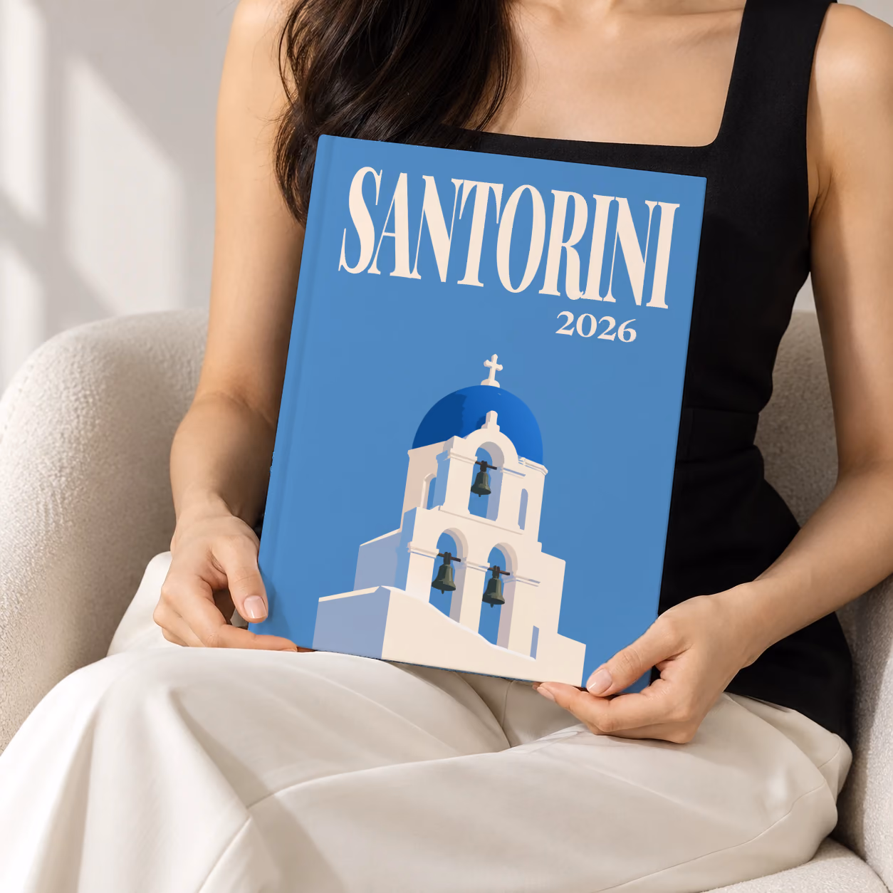 Greece hardcover photo book with the Santorini illustrated cover of a blue-domed bell tower on blue, held upright in two hands by a woman in a black top, sitting in a cream boucle armchair