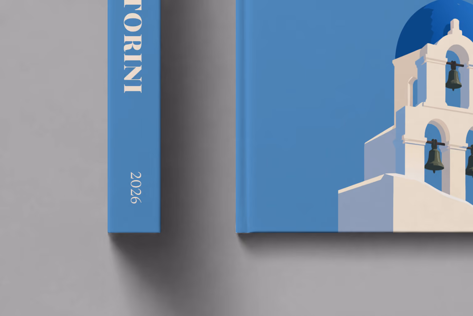 Greece hardcover photo book with the Santorini illustrated cover of a blue-domed bell tower on blue, shown close up beside its spine on a pale grey studio surface