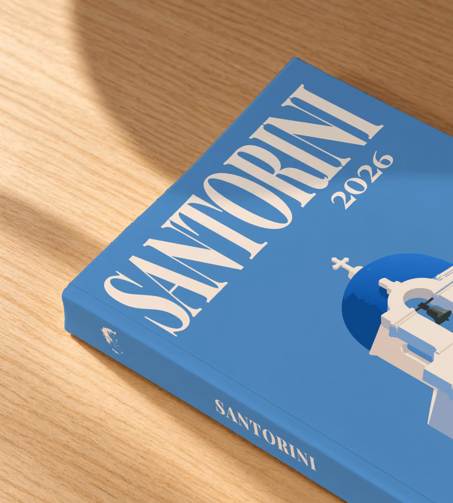Greece softcover photo book with the Santorini illustrated cover of a blue-domed bell tower on blue, lying on an oak desk in dappled afternoon sunlight