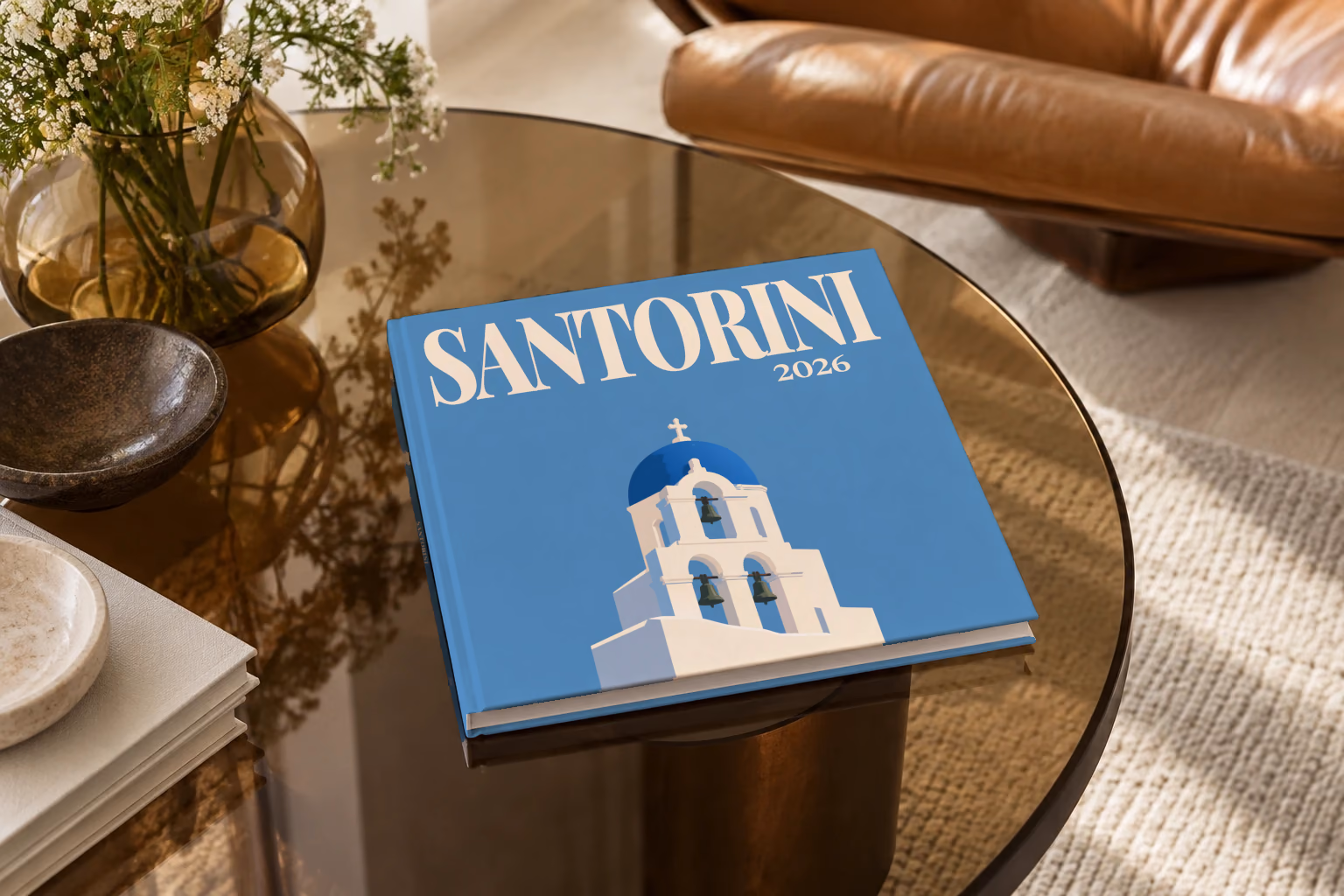 Greece square hardcover photo book with the Santorini illustrated cover of a blue-domed bell tower on blue, on a smoked glass coffee table beside a leather armchair in warm afternoon light