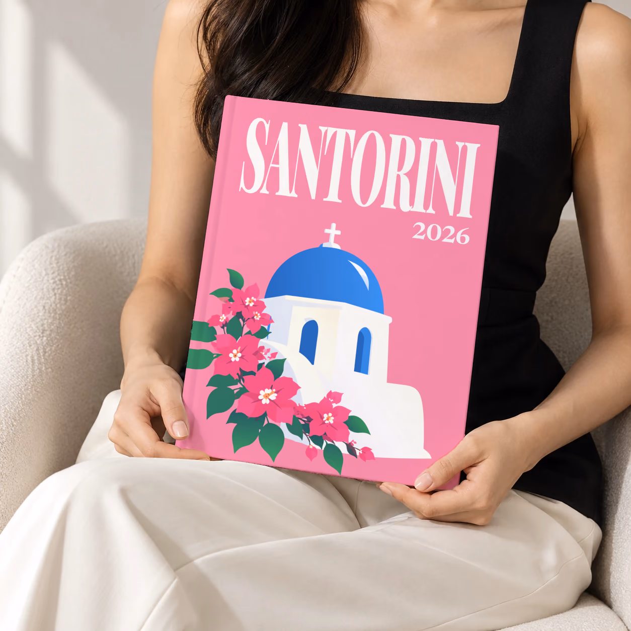 Greece hardcover photo book with the Santorini illustrated cover of a blue-domed chapel and bougainvillea on pink, held upright in two hands by a woman in a black top, sitting in a cream boucle armchair