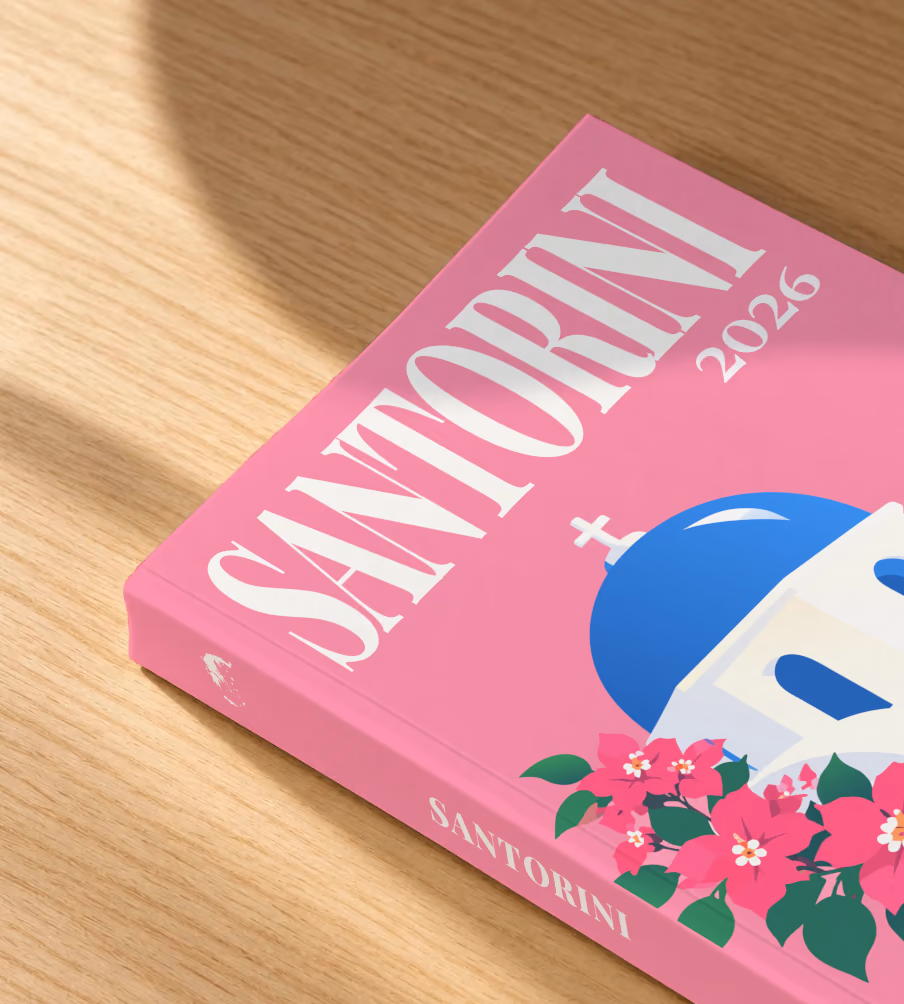 Greece softcover photo book with the Santorini illustrated cover of a blue-domed chapel and bougainvillea on pink, lying on an oak desk in dappled afternoon sunlight