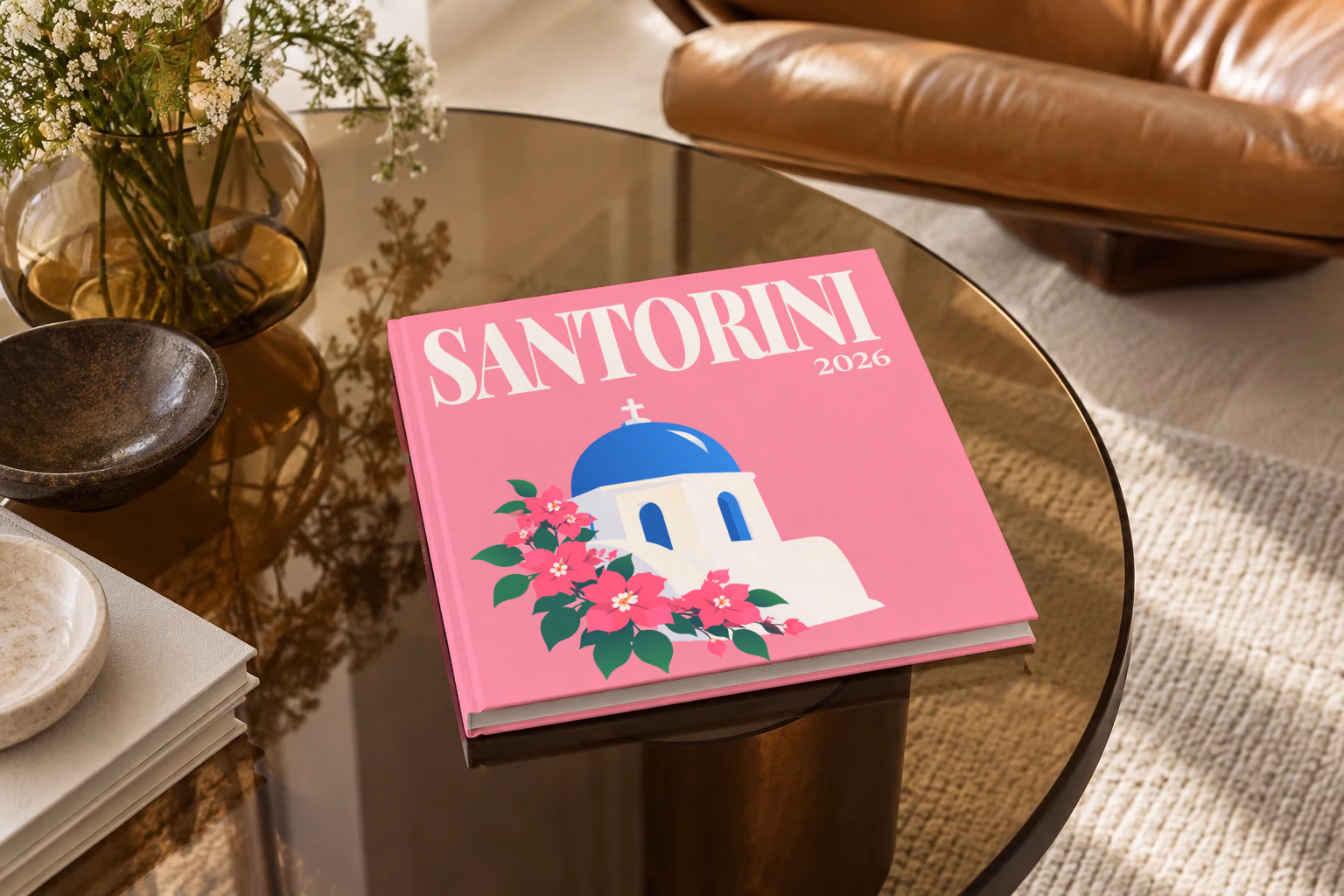 Greece square hardcover photo book with the Santorini illustrated cover of a blue-domed chapel and bougainvillea on pink, on a smoked glass coffee table beside a leather armchair in warm afternoon light