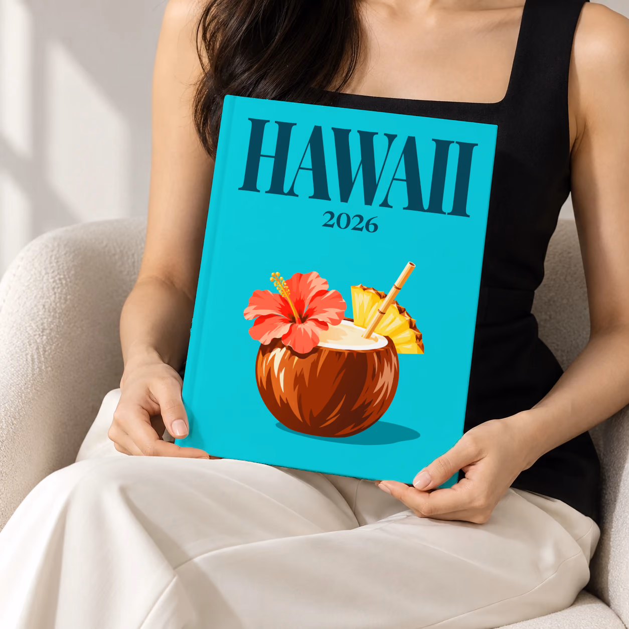 Hawaii hardcover photo book with the illustrated cover of a coconut drink with hibiscus and pineapple on turquoise, held upright in two hands by a woman in a black top, sitting in a cream boucle armchair