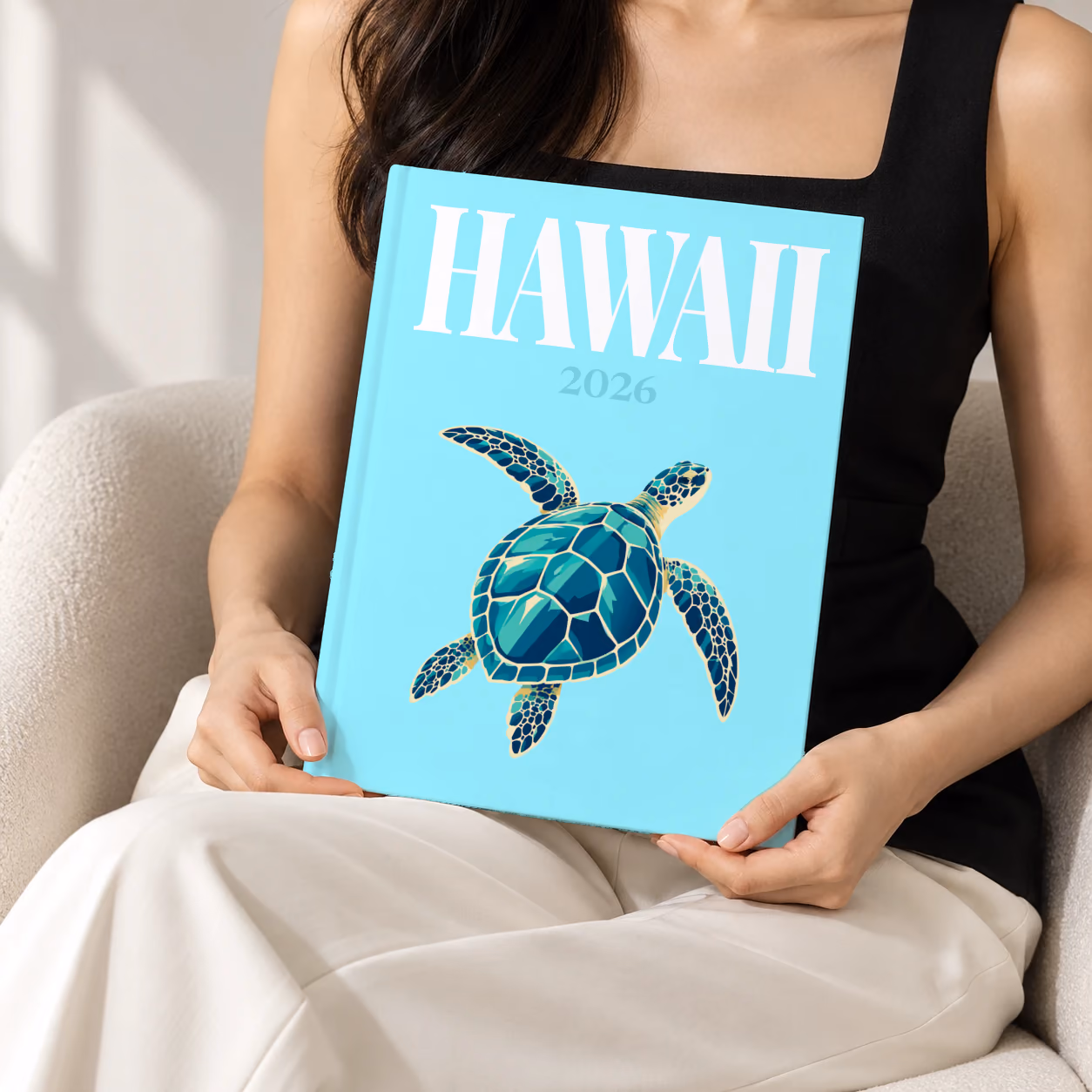 Hawaii hardcover photo book with the illustrated cover of a green sea turtle on pale aqua, held upright in two hands by a woman in a black top, sitting in a cream boucle armchair