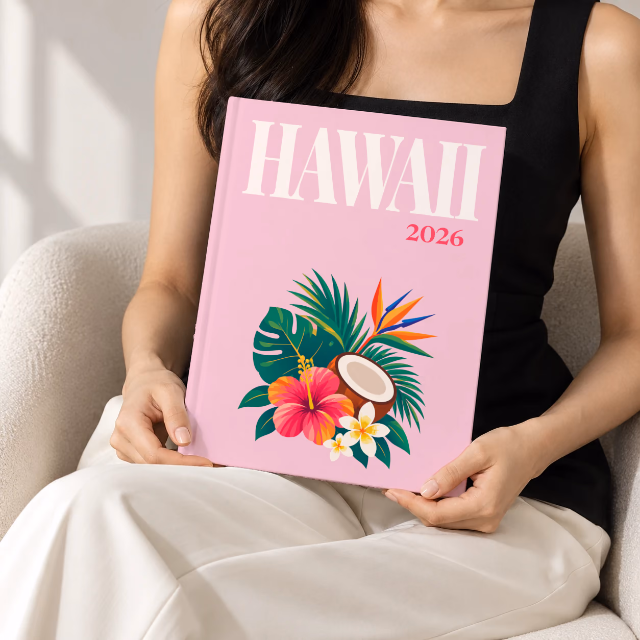 Hawaii hardcover photo book with the illustrated cover of a tropical bouquet with hibiscus, a coconut and palm leaves on pale pink, held upright in two hands by a woman in a black top, sitting in a cream boucle armchair