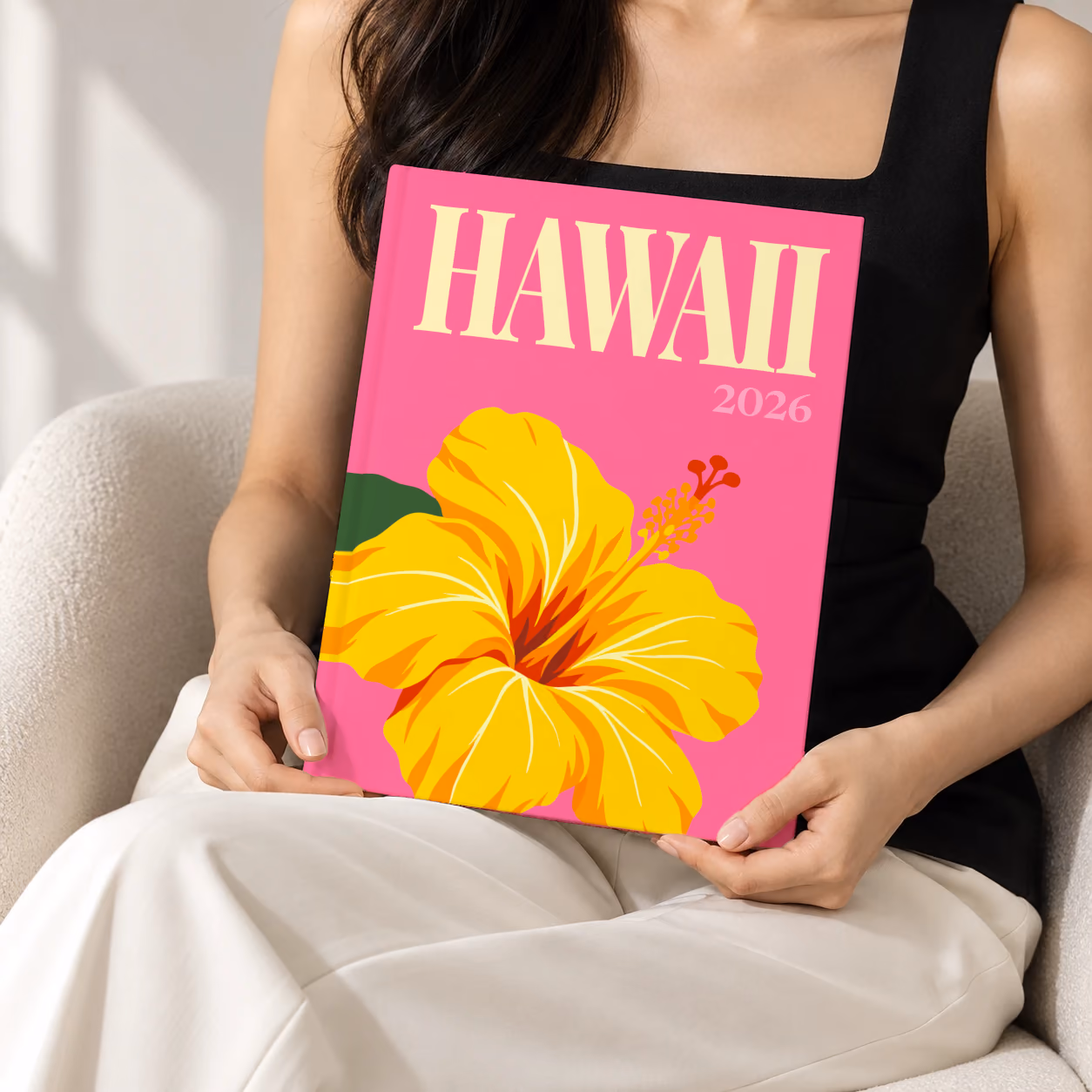 Hawaii hardcover photo book with the illustrated cover of a yellow hibiscus on pink, held upright in two hands by a woman in a black top, sitting in a cream boucle armchair