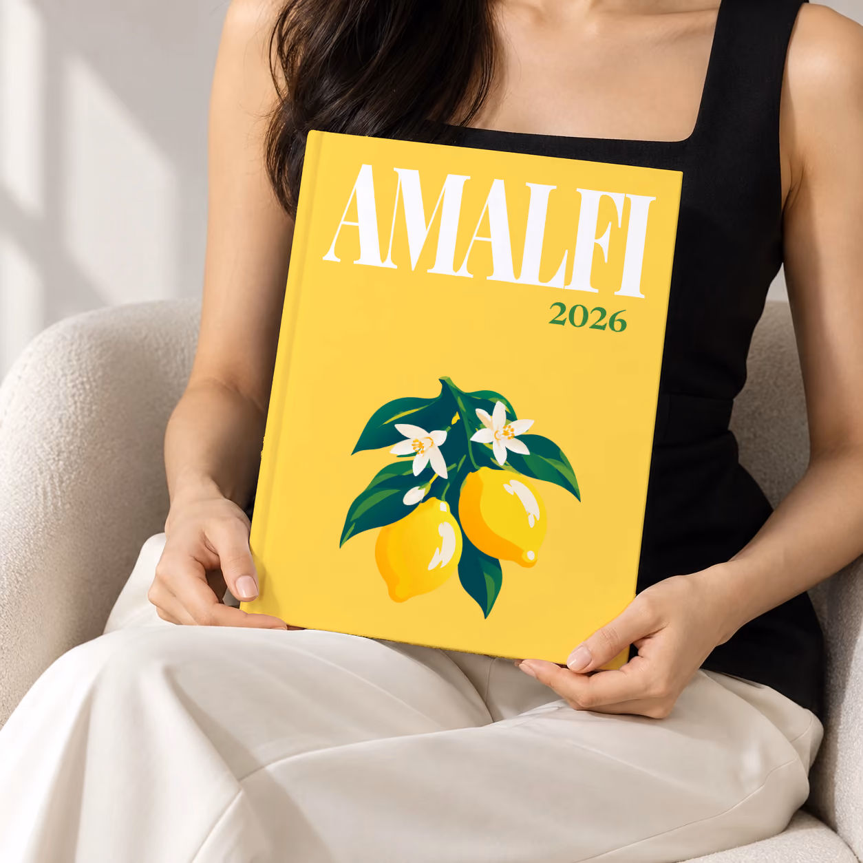 Italy hardcover photo book with the Amalfi illustrated cover of lemons and blossom on yellow, held upright in two hands by a woman in a black top, sitting in a cream boucle armchair