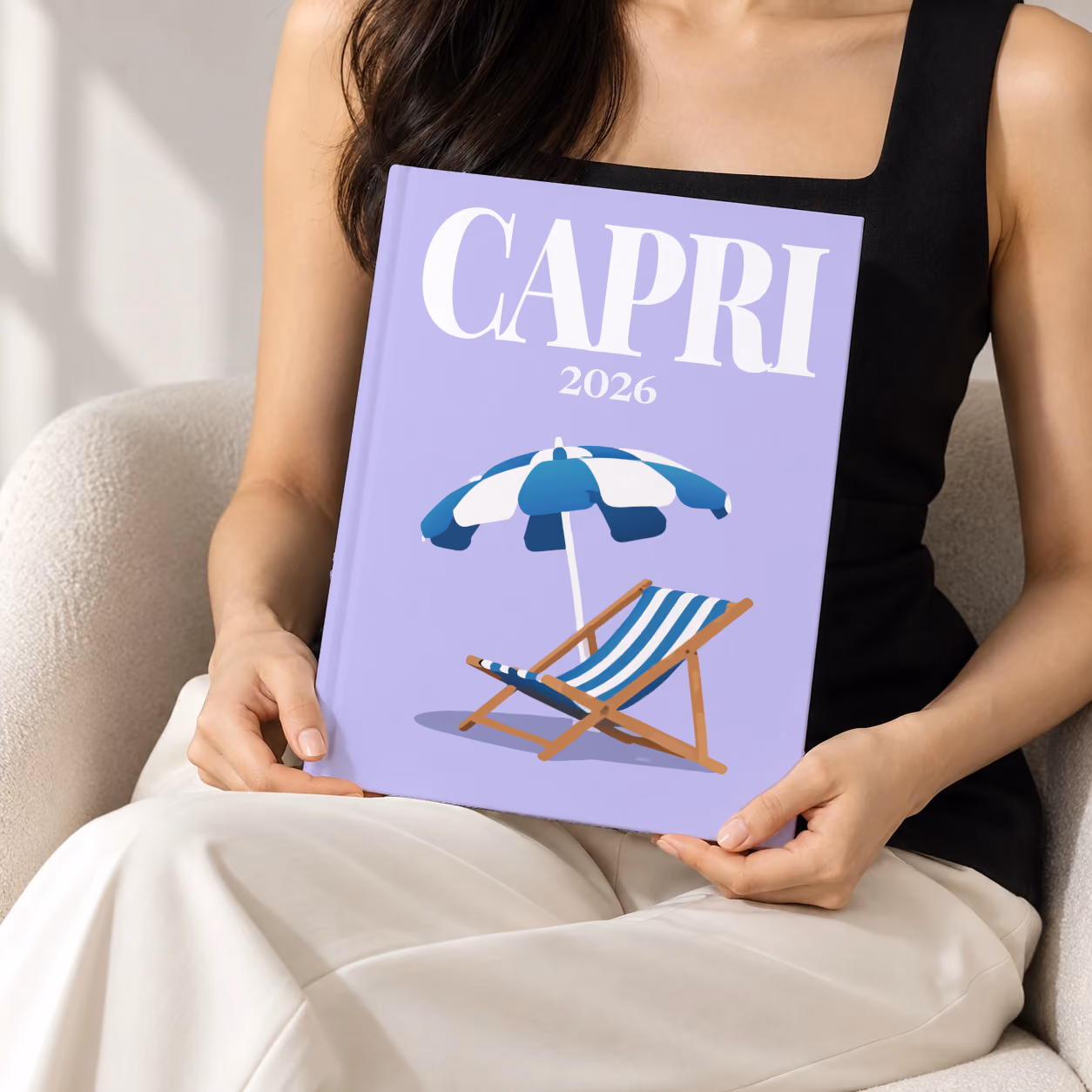 Italy hardcover photo book with the Capri illustrated cover of a striped beach umbrella and deckchair on lilac, held upright in two hands by a woman in a black top, sitting in a cream boucle armchair