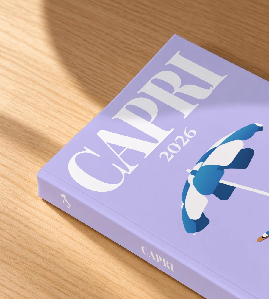 Italy softcover photo book with the Capri illustrated cover of a striped beach umbrella and deckchair on lilac, lying on an oak desk in dappled afternoon sunlight