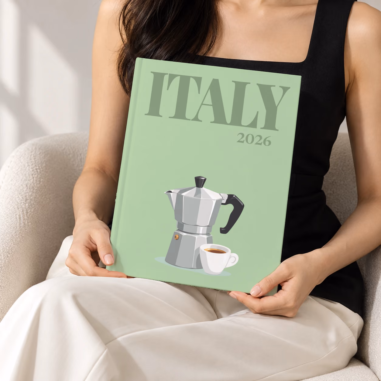 Italy hardcover photo book with the illustrated cover of a moka pot and an espresso cup on sage green, held upright in two hands by a woman in a black top, sitting in a cream boucle armchair