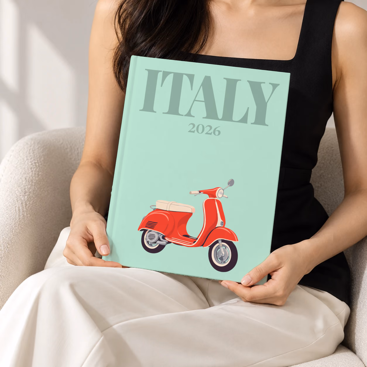 Italy hardcover photo book with the illustrated cover of a red Vespa scooter on mint green, held upright in two hands by a woman in a black top, sitting in a cream boucle armchair