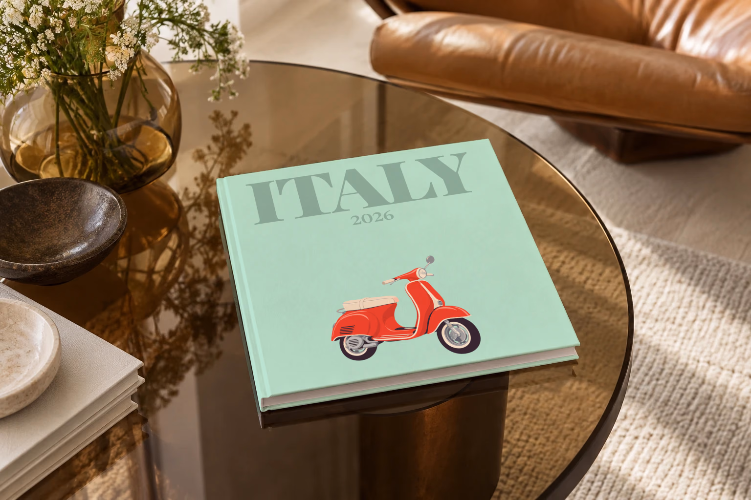 Italy square hardcover photo book with the illustrated cover of a red Vespa scooter on mint green, on a smoked glass coffee table beside a leather armchair in warm afternoon light