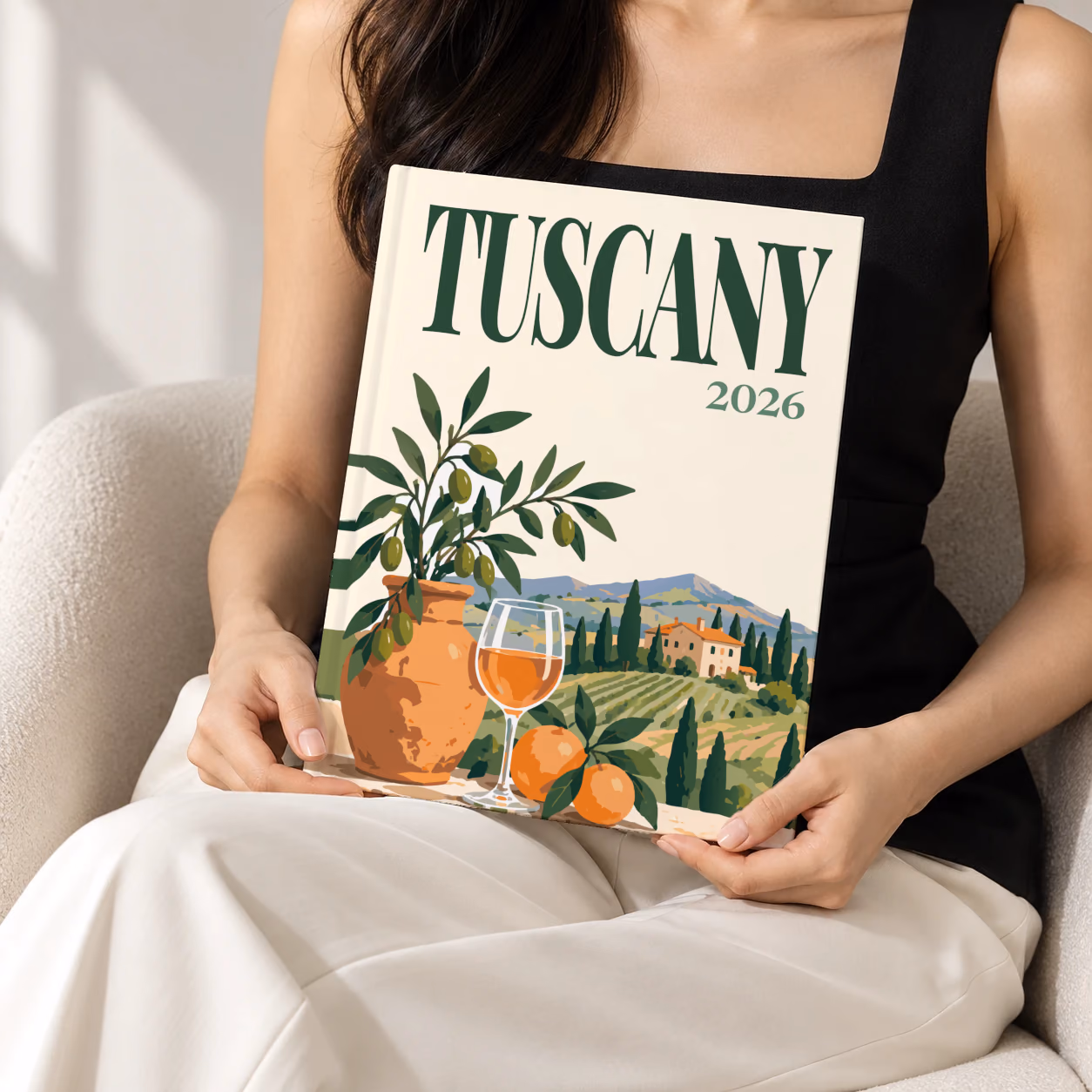 Italy hardcover photo book with the Tuscany illustrated cover of an olive tree, a glass of wine and Tuscan vineyards on cream, held upright in two hands by a woman in a black top, sitting in a cream boucle armchair