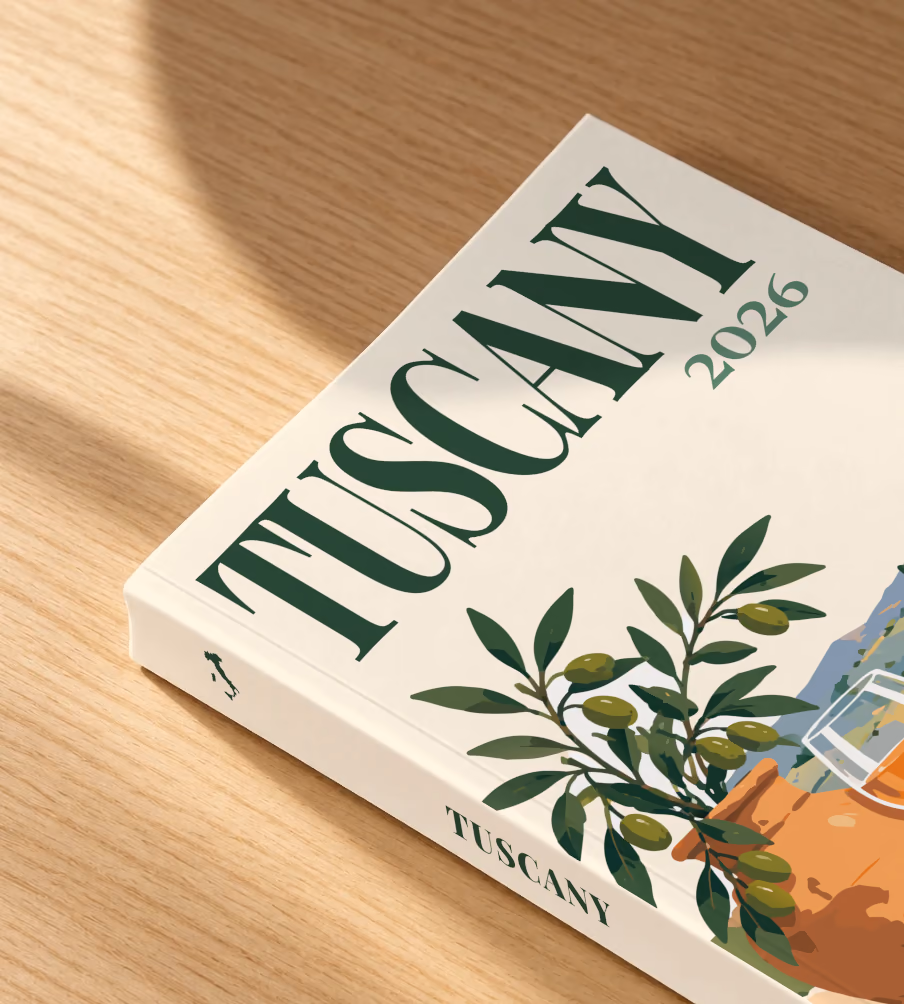 Italy softcover photo book with the Tuscany illustrated cover of an olive tree, a glass of wine and Tuscan vineyards on cream, lying on an oak desk in dappled afternoon sunlight