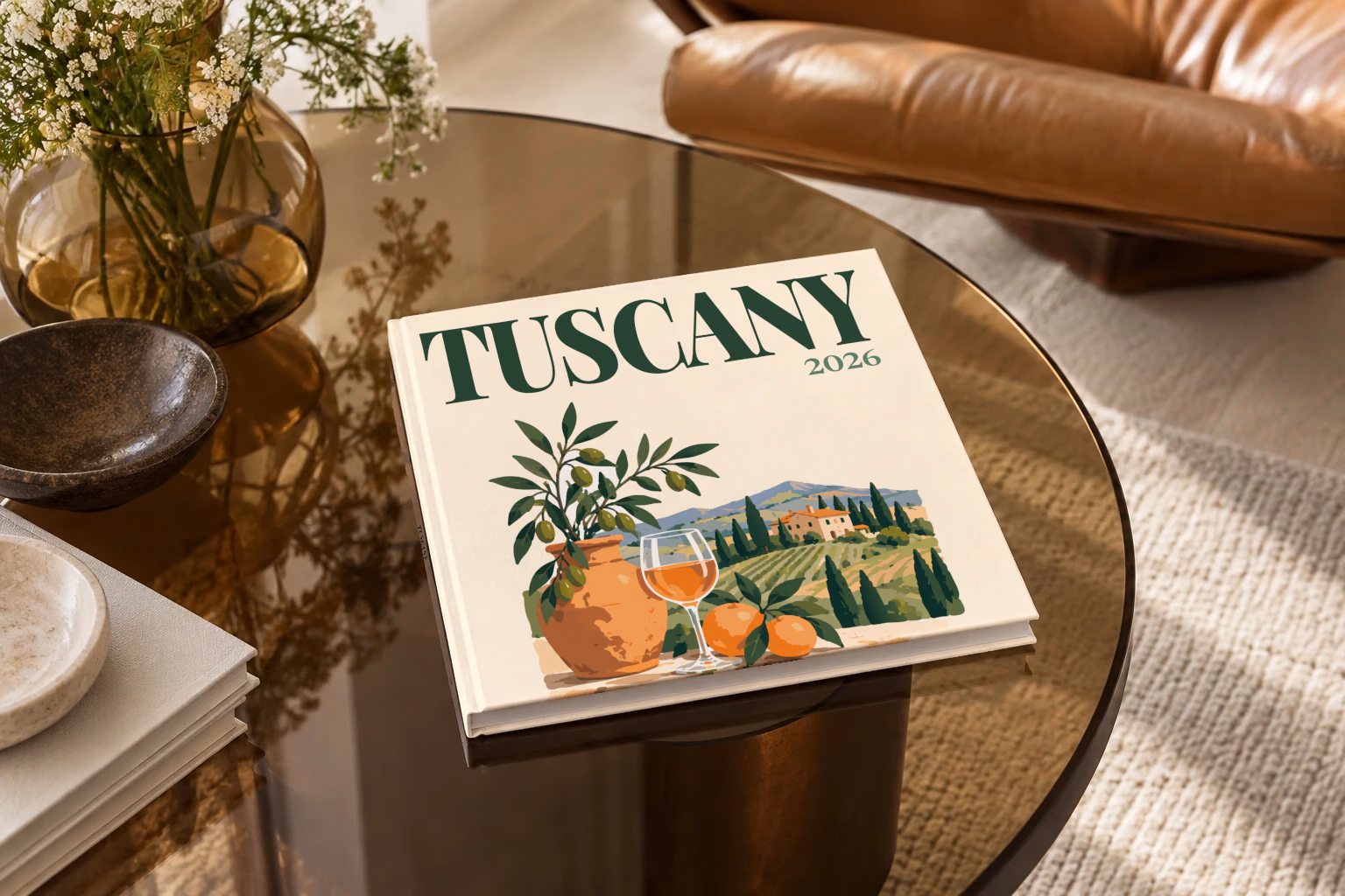 Italy square hardcover photo book with the Tuscany illustrated cover of an olive tree, a glass of wine and Tuscan vineyards on cream, on a smoked glass coffee table beside a leather armchair in warm afternoon light