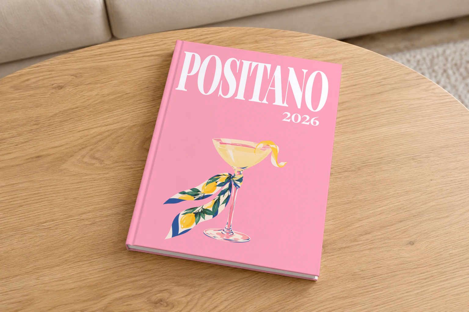 Italy hardcover photo book with the Positano illustrated cover of a martini glass tied with a lemon-print scarf on pink, on a round oak coffee table in a bright living room