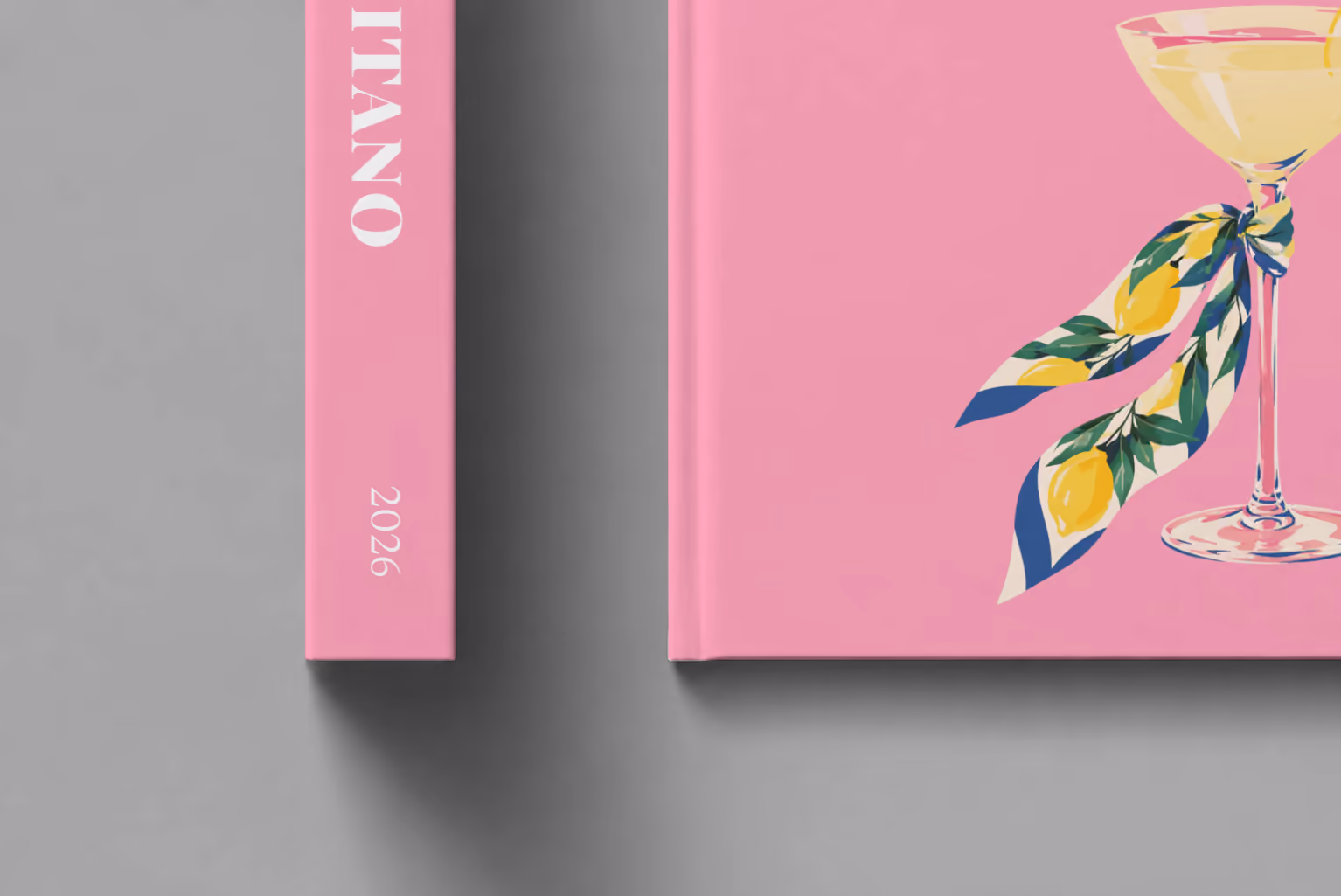 Italy hardcover photo book with the Positano illustrated cover of a martini glass tied with a lemon-print scarf on pink, shown close up beside its spine on a pale grey studio surface