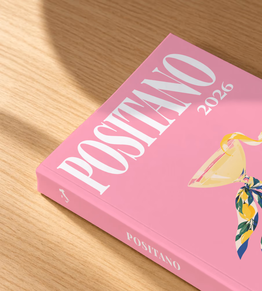 Italy softcover photo book with the Positano illustrated cover of a martini glass tied with a lemon-print scarf on pink, lying on an oak desk in dappled afternoon sunlight